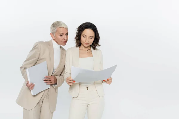 Young businesswoman holding paper folder near senior colleague isolated on grey — Stock Photo