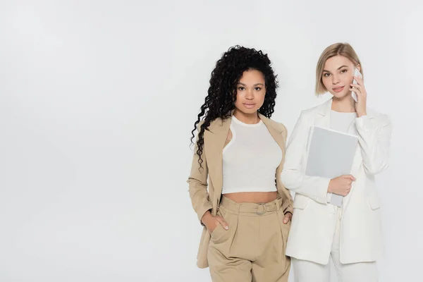 Interracial businesswomen with paper folder talking on smartphone and looking at camera isolated on grey — Stock Photo