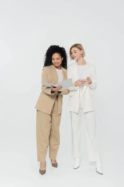 Positive african american businesswoman holding paper folder near colleague with smartphone on grey background — Stock Photo