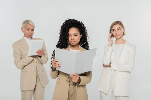 Donna d'affari afroamericana in possesso di cartella di carta mentre i colleghi utilizzano dispositivi isolati su grigio — Foto stock