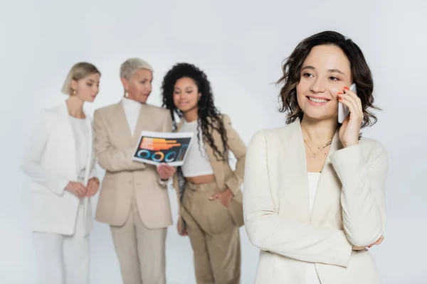 Femme d'affaires souriante parlant sur smartphone près de collègues multiethniques flous isolés sur gris — Photo de stock