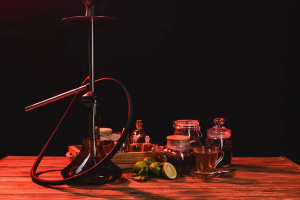 Thé, limes fraîches et narguilé sur table en bois isolé sur noir avec lumière rouge — Photo de stock