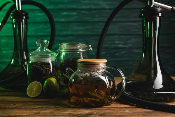 Teapot near blurred limes and hookahs on wooden table on wooden background — Stock Photo