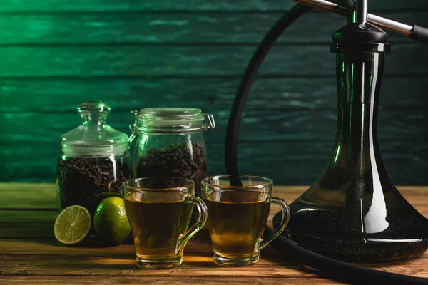 Cups of tea, limes and hookah on wooden surface on wooden background — Stock Photo