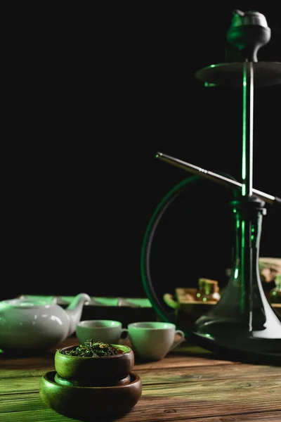Dry tea near blurred cups and hookah on wooden surface isolated on black — Stock Photo