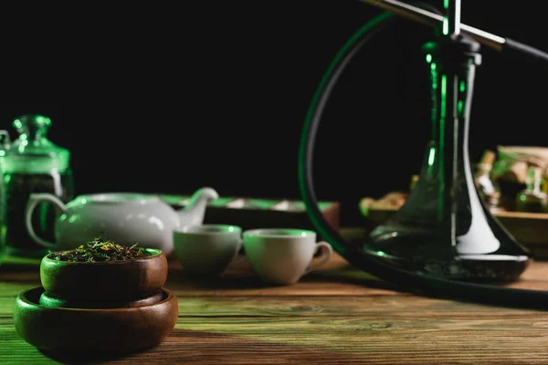 Chá seco perto de narguilé borrado e xícaras em mesa de madeira isolada em preto — Fotografia de Stock
