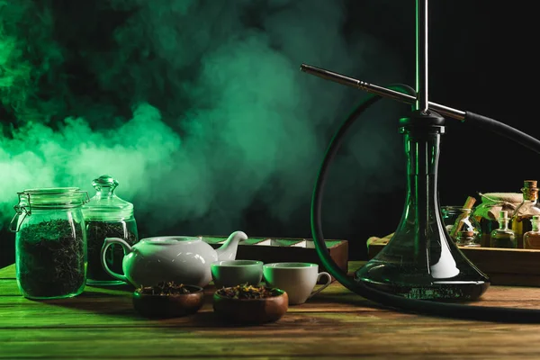 Hookah cerca de té seco y tetera en la superficie de madera sobre fondo negro con humo - foto de stock