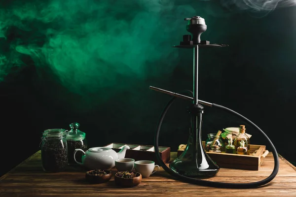 Chá e narguilé na mesa de madeira no fundo preto com fumaça — Fotografia de Stock