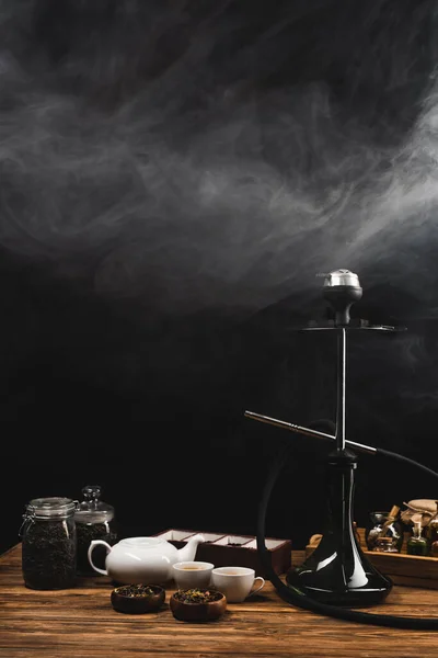 Tazas de té, hookah en la superficie de madera sobre fondo negro con humo - foto de stock