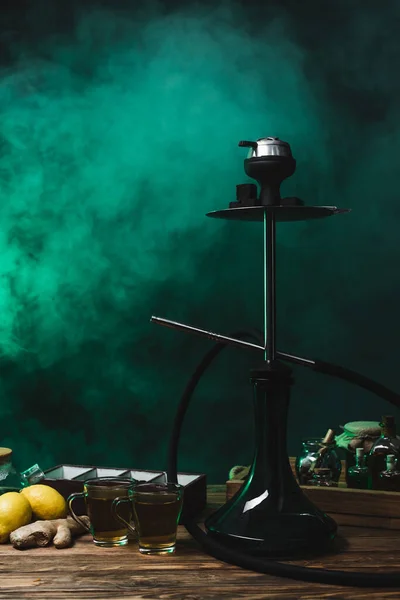 Hookah, lemons and tea on wooden surface on black background with smoke — Stock Photo