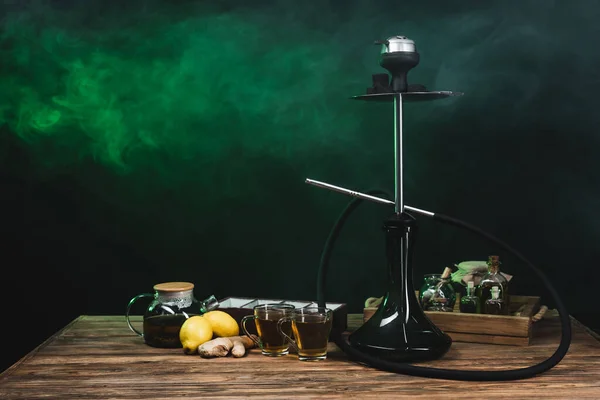 Lemons, tea and hookah on wooden surface on black background with smoke — Stock Photo