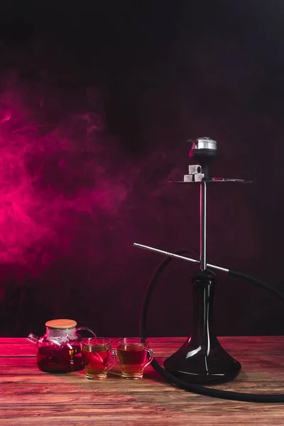 Hookah cerca de tazas de té en la superficie de madera sobre fondo negro con humo rosa - foto de stock