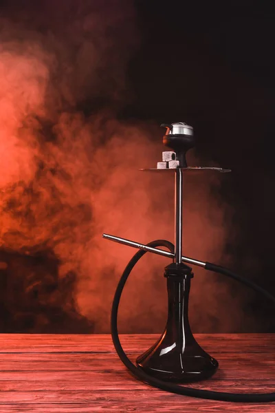 Hookah with coals on wooden surface on black background with red smoke — Stock Photo