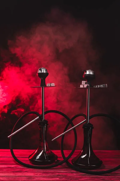 Hookahs avec des charbons sur la surface en bois sur fond noir avec de la fumée rouge — Photo de stock