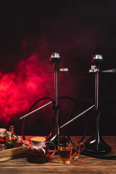 Cups of tea near teapot and hookahs on wooden surface on black background with red smoke — Stock Photo