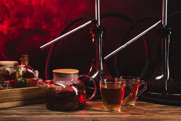 Tea near hookahs on wooden surface on black background with smoke — Stock Photo