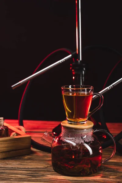 Cup of tea on teapot near hookah on wooden surface isolated on black — Stock Photo