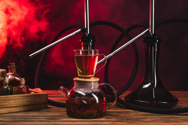 Cup of tea on pot near hookahs on wooden surface on black background with red smoke — Stock Photo