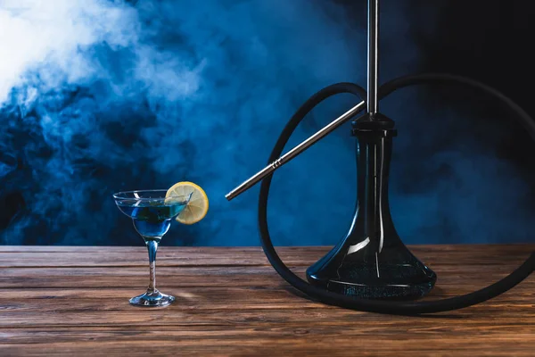 Glass with cocktail near hookah on wooden surface on black background with smoke — Stock Photo