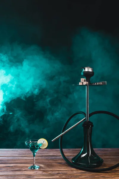 Hookah cerca de vaso de cóctel en la superficie de madera sobre fondo negro con humo verde - foto de stock