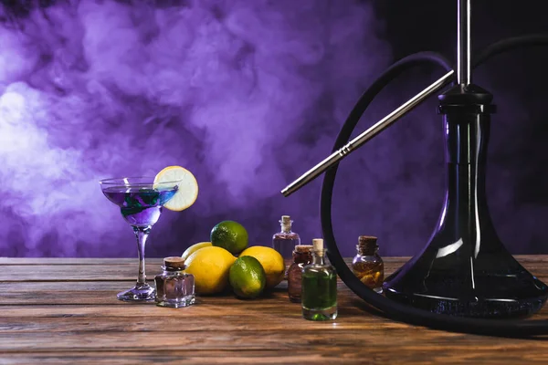 Hookah near fruits and cocktail on black background with purple smoke — Stock Photo