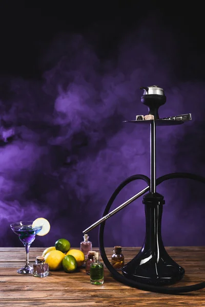 Hookah near cocktail on wooden surface on black background with purple smoke — Stock Photo