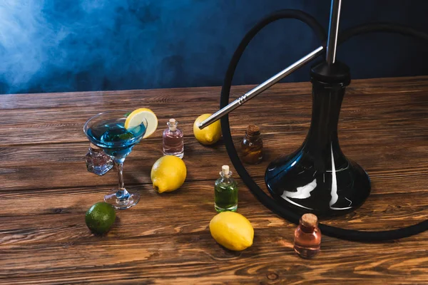 Hookah near lemons and glass of cocktail on wooden surface on black background with blue smoke — Stock Photo