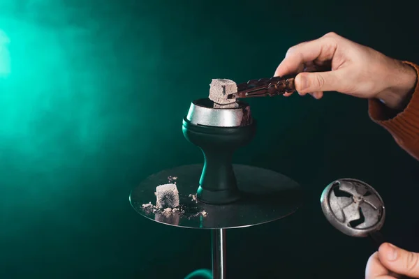 Cropped view of man holding coal in tweezers near hookah on green background with smoke — Stock Photo