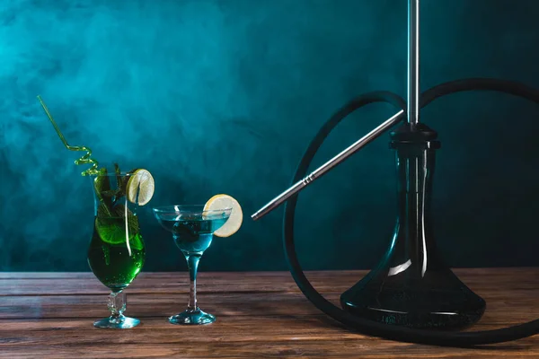 Glass of cocktails near hookah on wooden surface on black background with green smoke — Stock Photo