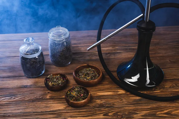 Dry tea in jars and bowls near hookah on wooden surface on black background with smoke — Stock Photo