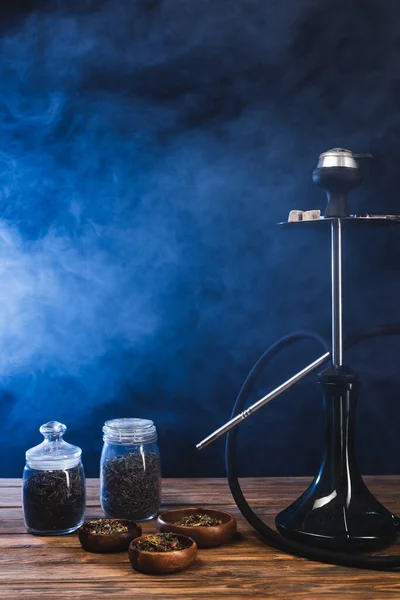 Hookah near dry tea in bowls on wooden surface on black background with blue smoke — Stock Photo