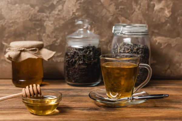 Cup of tea near honey and blurred jars on wooden surface on textured stone background — Stock Photo