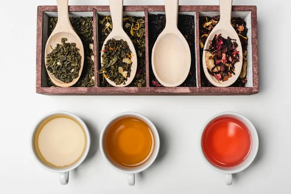 Vue du dessus du thé dans des tasses près de cuillères en bois et boîte sur fond blanc — Photo de stock