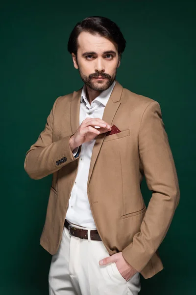 Jeune homme avec moustache ajustant mouchoir dans poche de blazer beige sur vert foncé — Photo de stock