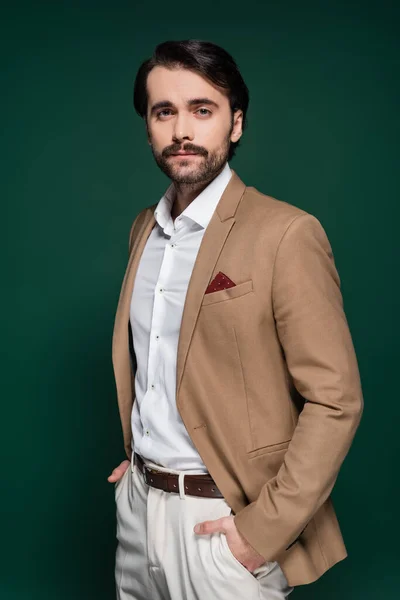 Young man with mustache looking at camera and posing with hands in pockets on dark green — Stock Photo