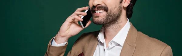 Vista recortada de joven feliz con bigote hablando en el teléfono inteligente en verde oscuro, bandera - foto de stock