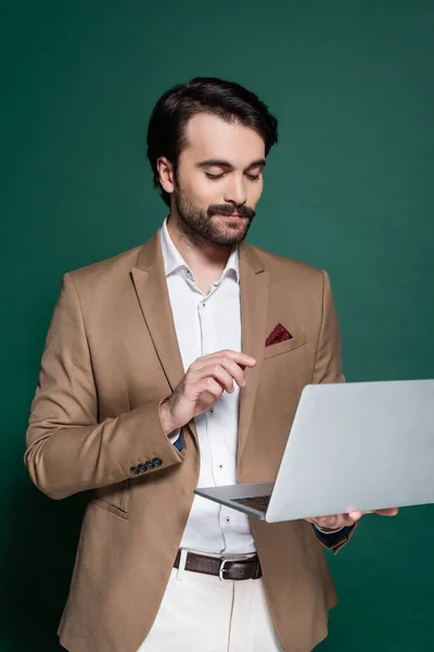 Jovem satisfeito com bigode usando laptop em verde escuro — Fotografia de Stock