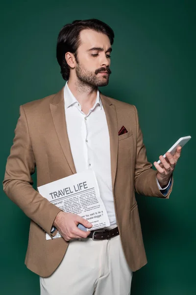 Joven con bigote sosteniendo periódico de viaje y el uso de teléfono inteligente en verde oscuro — Stock Photo
