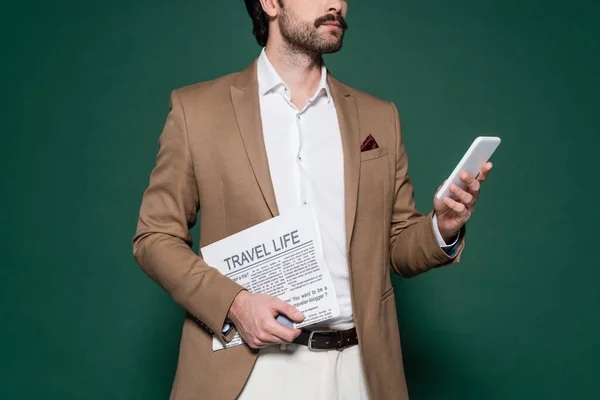 Cropped view of young man with mustache holding travel newspaper and using smartphone on dark green — Stock Photo