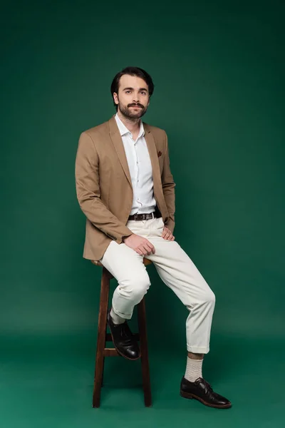 Pleine longueur de l'homme avec moustache assis sur une chaise en bois sur vert foncé — Photo de stock