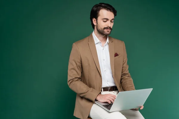 Man with mustache sitting on wooden chair and using laptop on dark green — Stock Photo