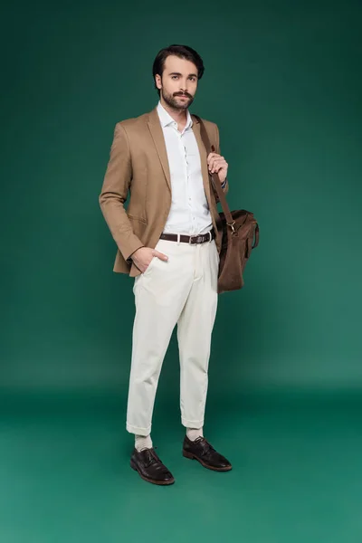 Мужчина в полный рост с горсткой стоит с рукой в кармане и держит кожаную сумку на темно-зеленом — стоковое фото
