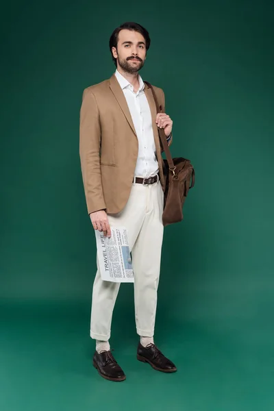 Full length of man with mustache holding travel newspaper and standing with leather bag on dark green — Stock Photo