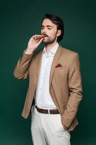 Homme avec moustache fumant cigare et debout avec la main dans la poche sur vert foncé — Photo de stock