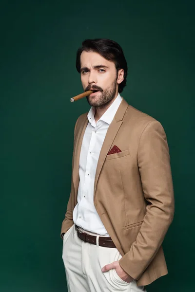 Man with mustache smoking cigar and standing with hands in pockets on dark green — Stock Photo