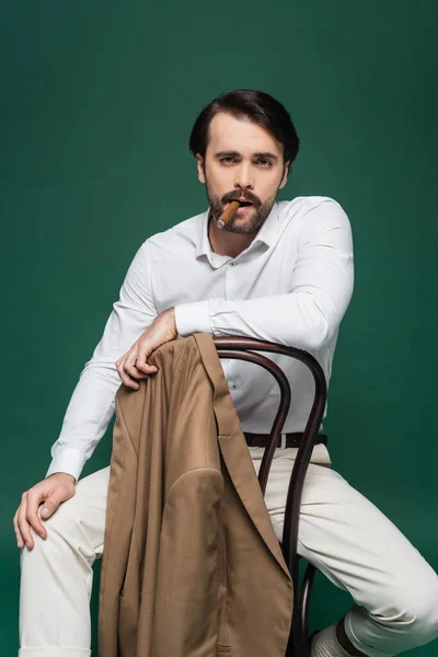 Man with mustache smoking cigar and sitting on wooden chair on dark green — Stock Photo