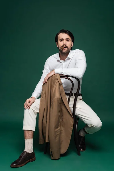 Full length of man with mustache smoking cigar and sitting on wooden chair on dark green — Stock Photo