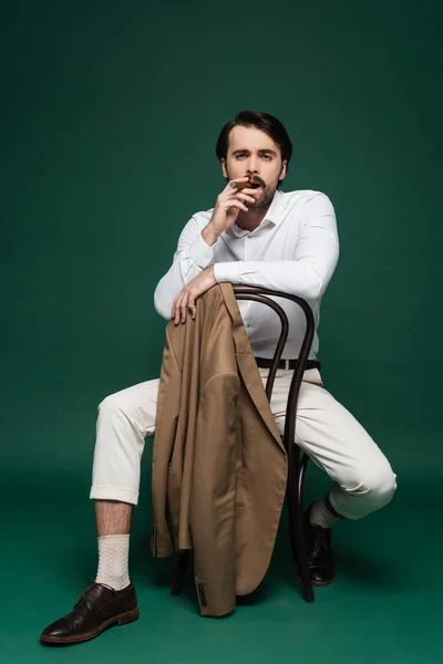 Pleine longueur de l'homme avec moustache fumant cigare et assis sur la chaise sur vert foncé — Photo de stock