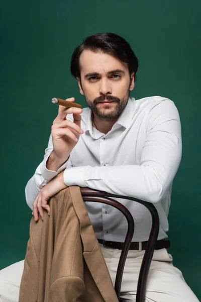 Homme avec moustache tenant cigare et assis sur la chaise sur vert foncé — Photo de stock