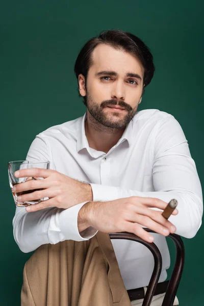 Hombre con bigote celebración cigarro y vaso de whisky mientras se apoya en la silla en verde oscuro - foto de stock